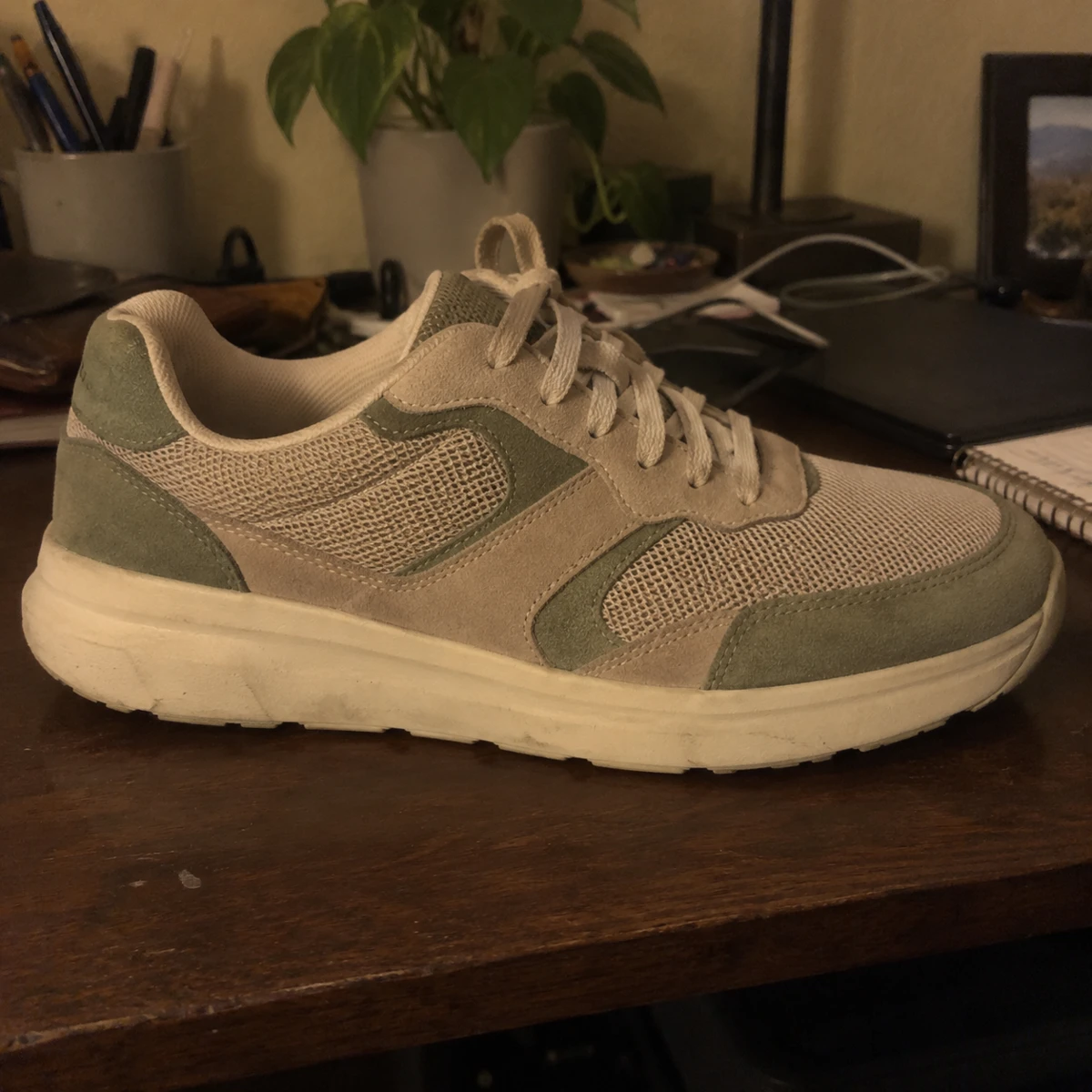 Illustrative quick phone photograph of a beige and sage sneaker in dim yellow light on a cluttered desk